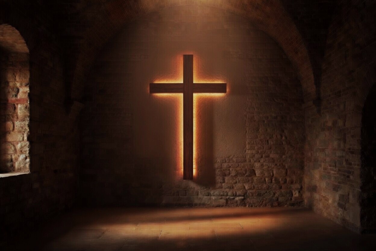 Glowing cross mounted on a stone wall in a dim, vaulted interior. Light softly outlines the cross from behind.